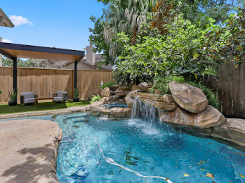 17839042952687853455835486363304 - Sage Pools, Scapes & Design Backyard pool with a rock waterfall feature, clear blue water, and a shaded seating area under a pergola.
