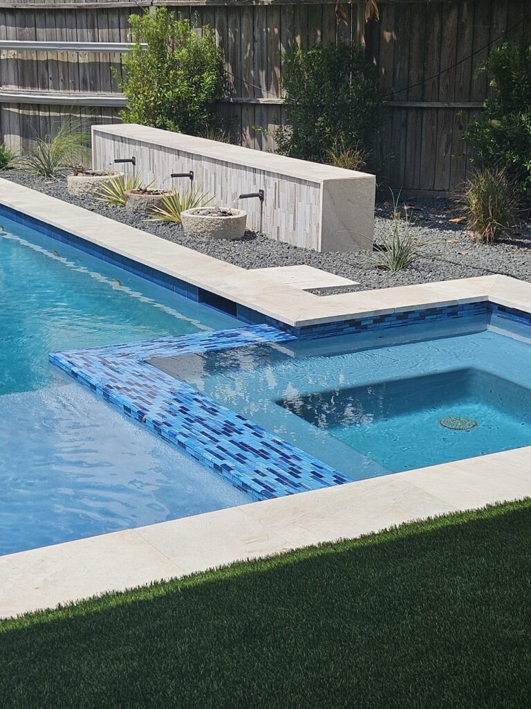 20260915_130913 - Sage Pools, Scapes & Design Backyard pool with built-in water spouts spraying into the blue-tiled edge, surrounded by gravel and planters with desert plants and a wooden fence in the background.