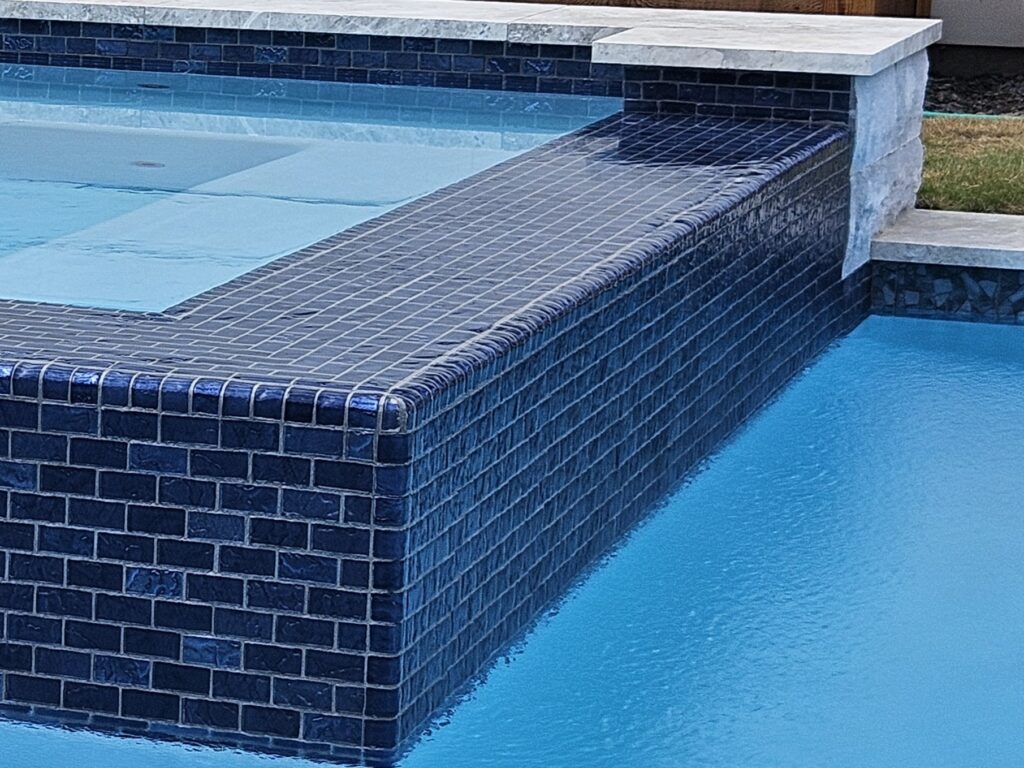 20261019_161430 - Sage Pools, Scapes & Design Close-up of a dark blue tiled pool edge with light stone coping visible above the tiles.