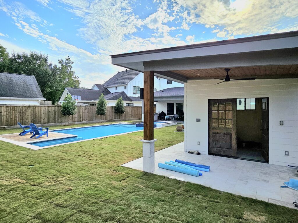 20261019_161536 - Sage Pools, Scapes & Design Backyard with a rectangular blue swimming pool, blue lounge chairs, and a white-tiled patio next to a modern house under a partly cloudy sky.